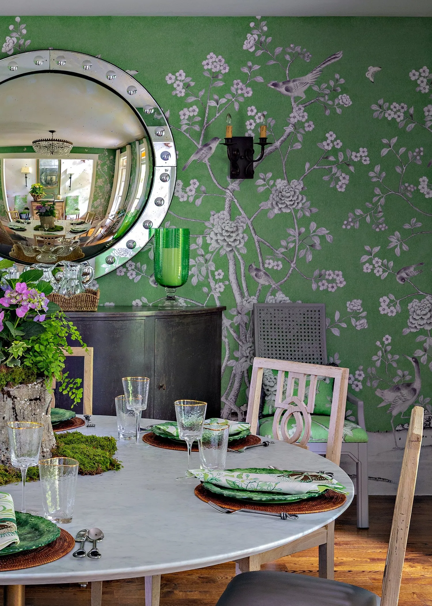 A dining room papered in green chinoiserie, a studded convex mirror over a dark sideboard and a marble table laid for lunch.
