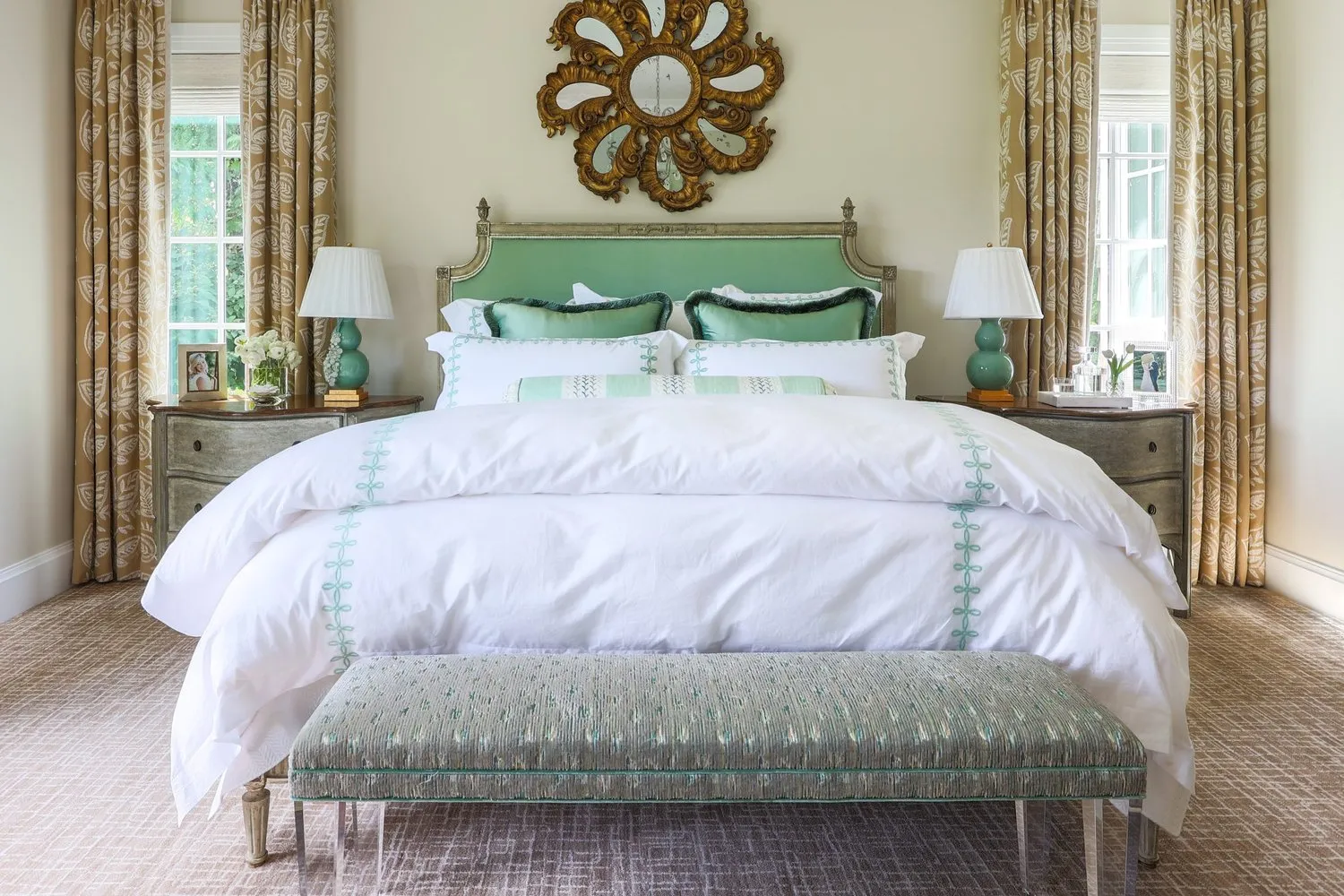 A primary bedroom with a green painted headboard centred under a carved gilt mirror, matching lamps and chests either side.