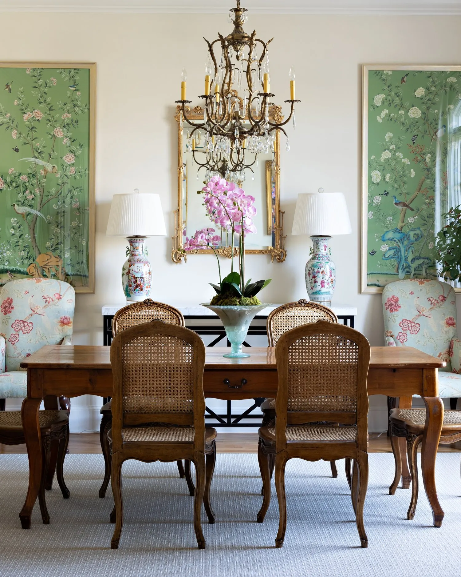 A dining room hung with a pair of green chinoiserie panels either side of a gilt mirror, a crystal chandelier over a fruitwood table.