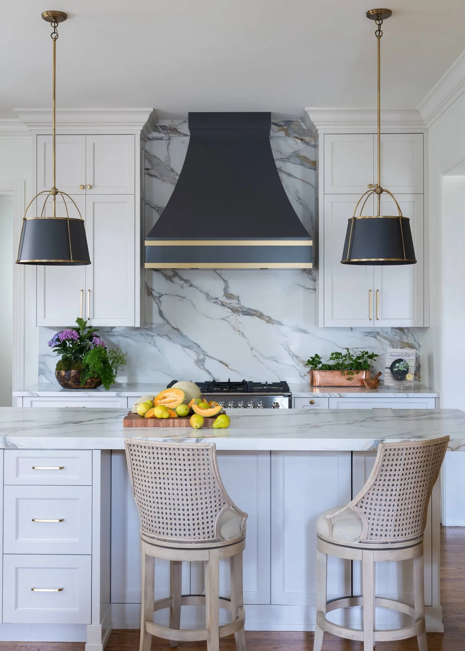 A white kitchen with a marble slab backsplash under a navy hood banded in brass, a pair of black shaded pendants over the island.